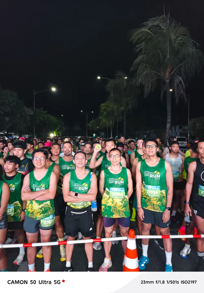 A group of male runners in green race singlets standing together behind a barrier at night, looking focused and ready for the event to commence.