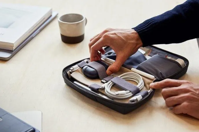 A close-up of a person’s hands organizing the interior of a charcoal Bellroy Tech Kit. The pouch is open, revealing neatly arranged cables, a power bank, and small tech accessories held in place by elastic loops and pockets.