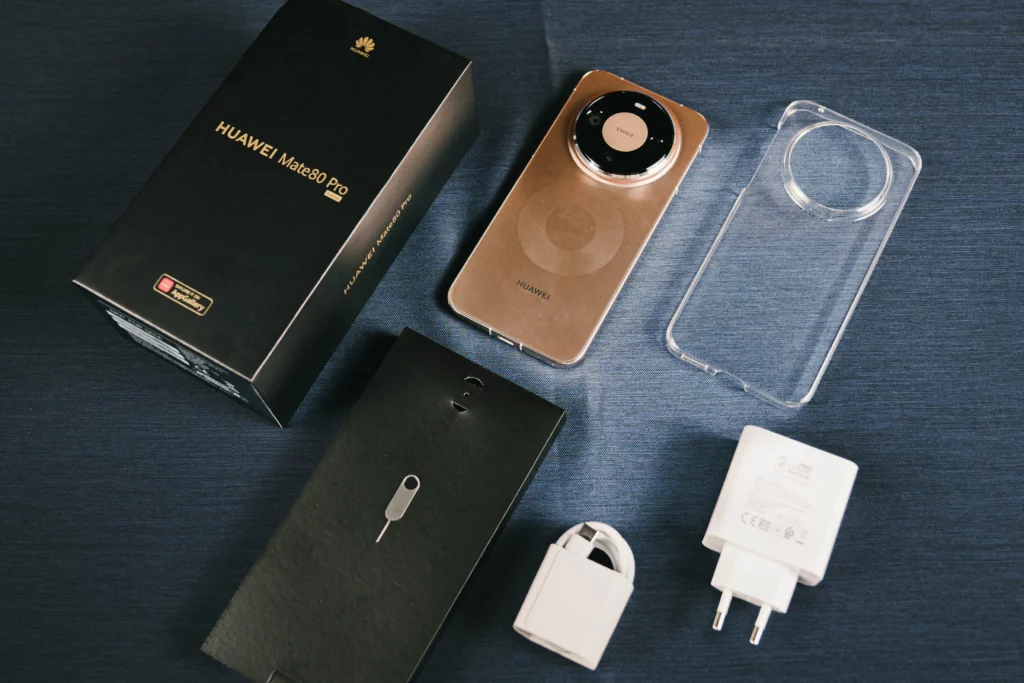 An unboxing layout on a blue textured surface. Contents include the HUAWEI Mate 80 Pro handset, a clear protective case, a white SuperCharge power adapter, a USB-C cable, and a SIM ejector tool pinned to a black cardboard sleeve.