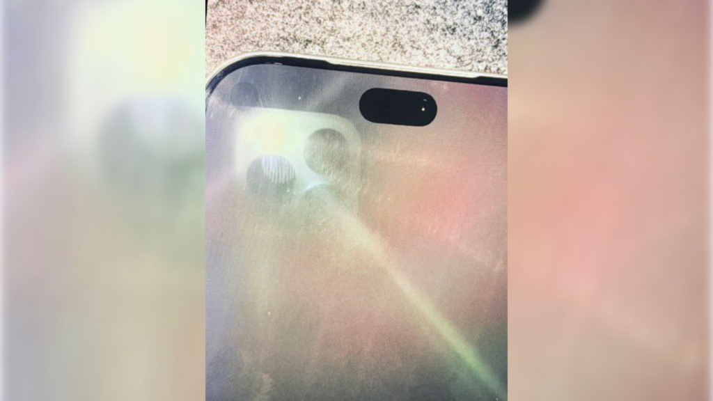 A close-up, slightly blurry photograph of a smartphone's upper display, purportedly an iPhone 18 Pro prototype. A bright light is being shone on the screen, revealing a small, circular punch-hole camera lens hidden beneath the glass, situated next to a significantly narrowed pill-shaped Dynamic Island cutout.