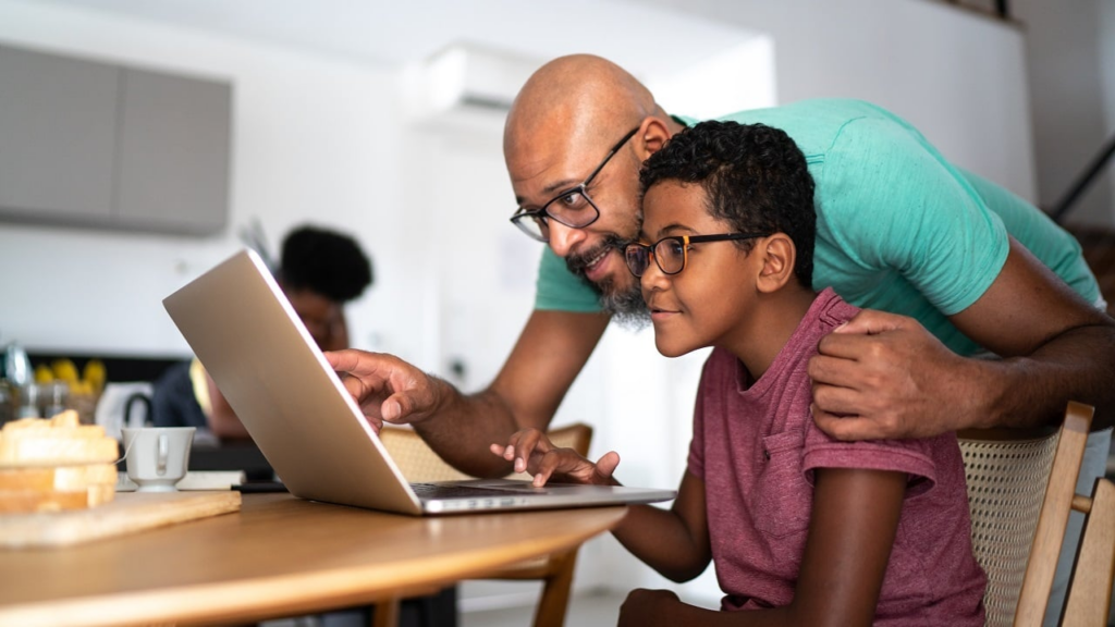 A father with glasses and a beard leaning over a wooden table to help his young son use a silver laptop. The father has his hand on the boy's shoulder in a supportive gesture while pointing at the screen in a bright, modern home setting.