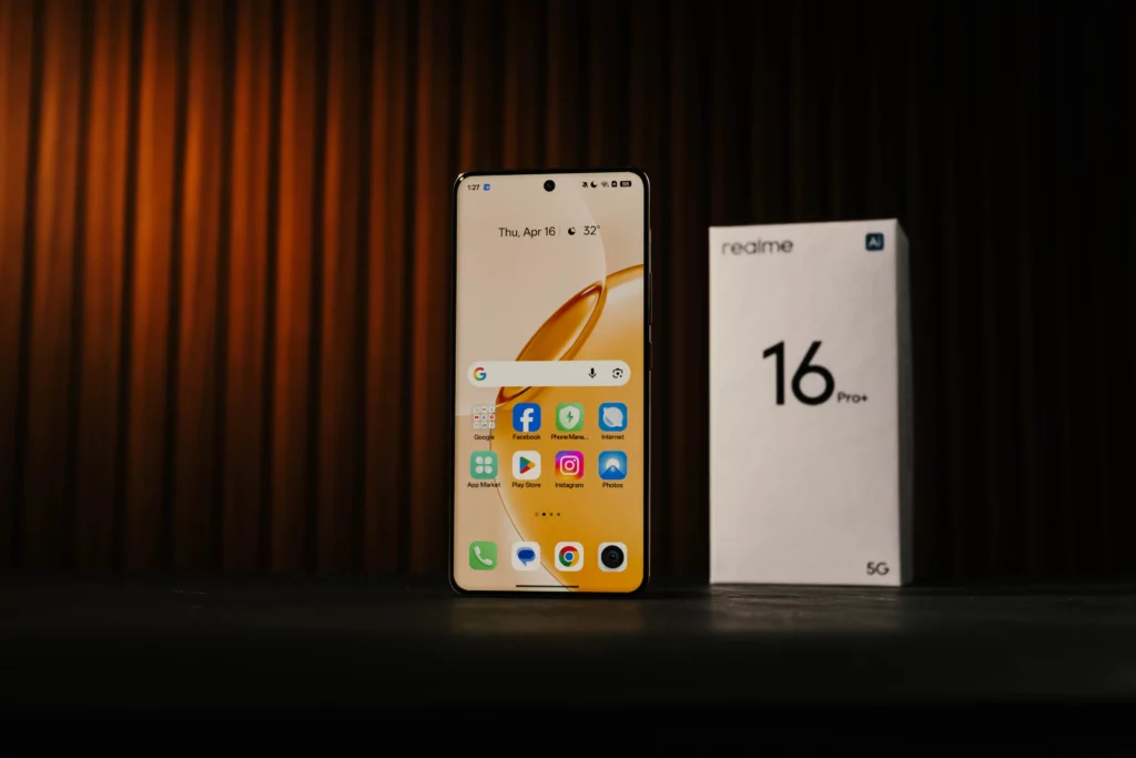 The realme 16 Pro+ 5G standing vertically with its screen showing the home page while the gray box stands behind it vertically.