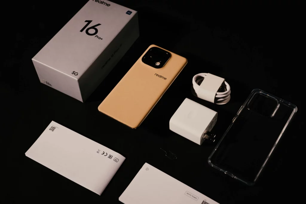 The realme 16 Pro+ 5G's contents laid out on a dark table showing the box, the phone itself, a charging brick, a charging cable, the case, and some documents at the bottom.