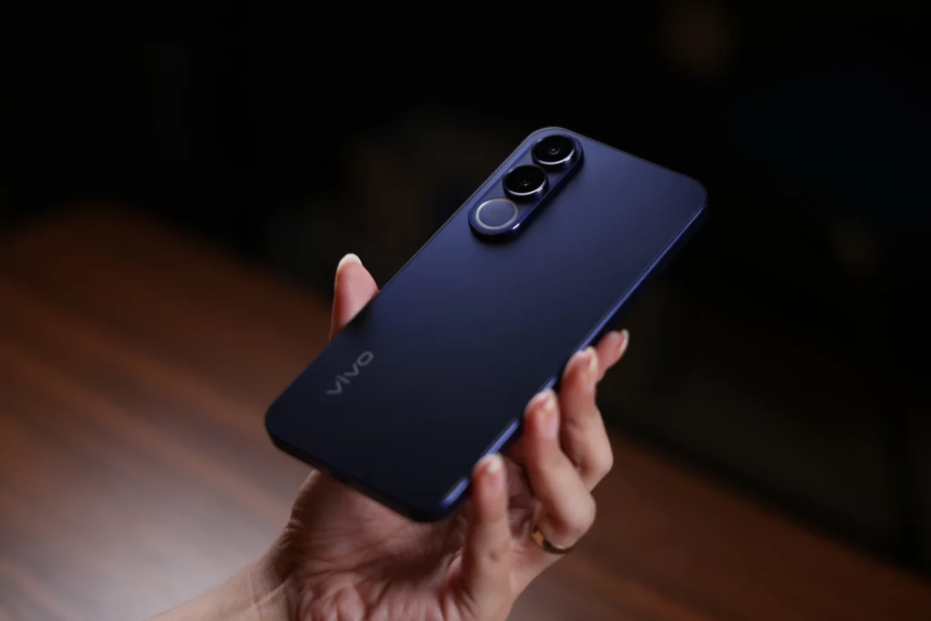 A person’s hand holding the vivo V70 FE at an angle, showcasing the smooth, matte purple finish and the prominent triple-camera module on the back. The lighting creates a soft gradient across the phone’s surface.