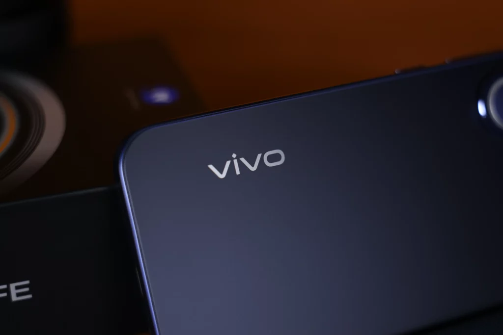 A close-up shot focusing on the silver "vivo" logo on the lower back of the matte purple handset. The retail box is partially visible in the blurred background.
