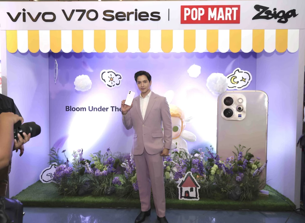 Alden Richards stands in a themed "Bloom Under The Stars" pop-up booth, holding a white vivo V70 FE smartphone. The booth features floral decorations, a large model of a smartphone camera module, and POP MART Zsiga branding.