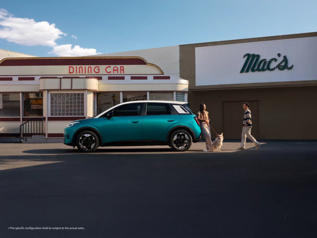 A side profile view of the compact GAC AION UT EV in an Emerald Green and Rococo White two-tone color scheme. The car is parked on an asphalt lot in front of retro-style buildings, including a diner labeled Dining Car and a storefront named Mac's, while a woman walks a dog nearby.