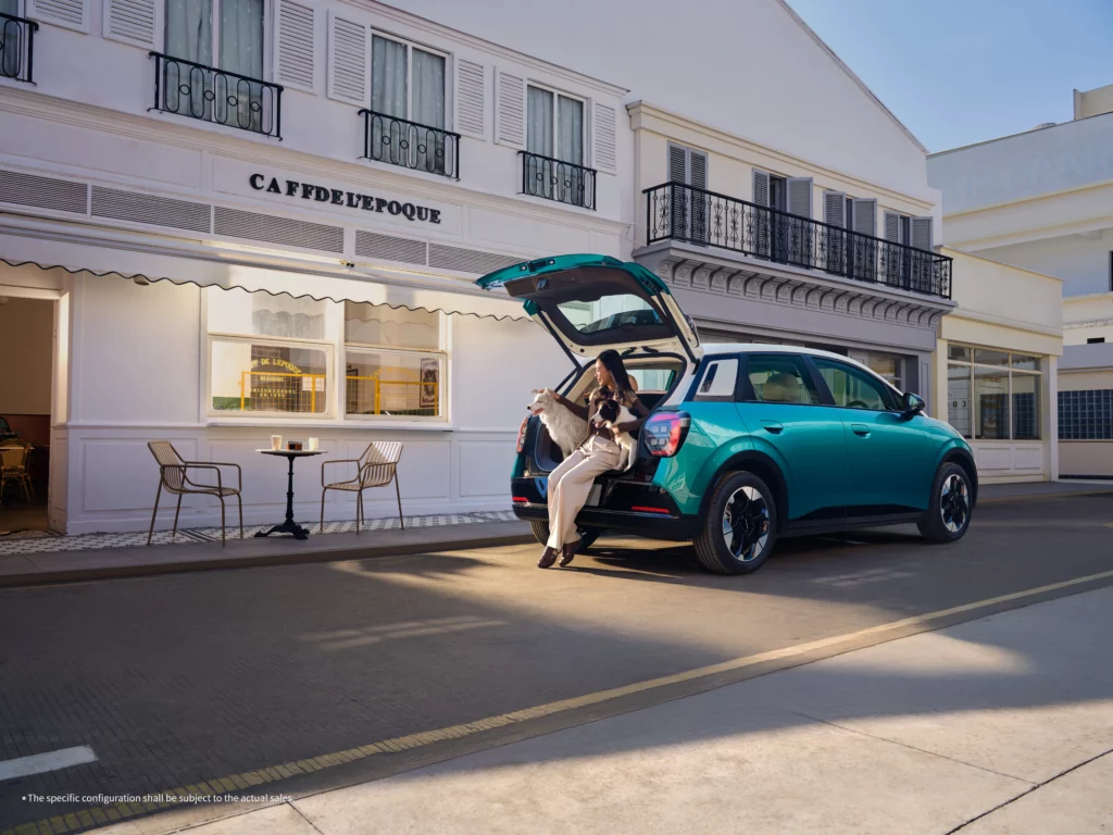 A teal and white two-tone GAC AION UT hatchback parked on a sunny city street in front of a European-style white building named Caffd' L'Epoque. The rear tailgate is open, and a woman is sitting in the spacious cargo area with two fluffy dogs, showcasing the vehicle's practical urban utility.