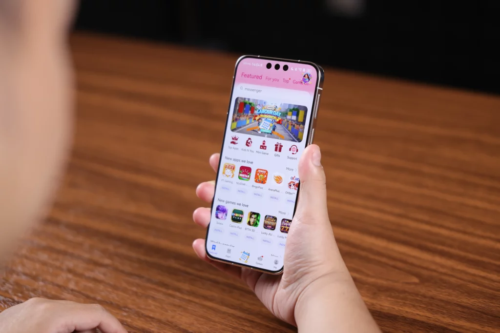 A close-up shot of a person holding a gold-trimmed smartphone showing the Huawei AppGallery home screen. The interface displays "Featured" and "New apps we love" categories with various app icons. A finger is near the bottom edge of the screen, and a wooden table surface is visible in the background.