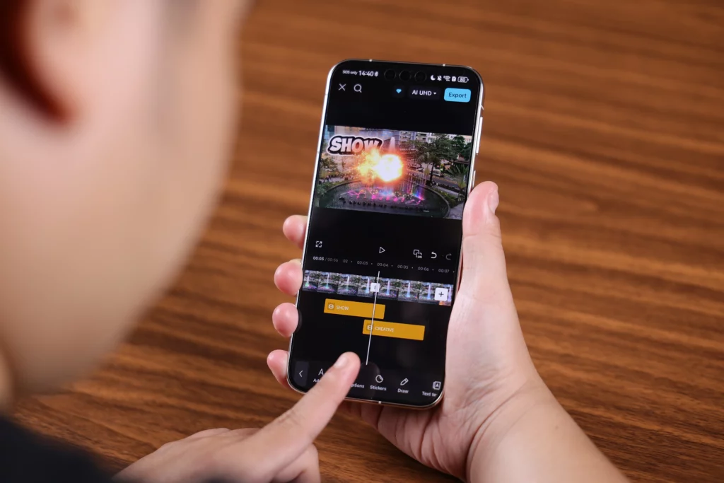 A close-up over-the-shoulder view of a person using an index finger to edit a video on a smartphone. The video editing timeline shows multi-layer tracks with yellow text blocks, a video preview featuring a colorful fountain display with a fire effect overlay, and an "AI UHD" export toggle at the top.