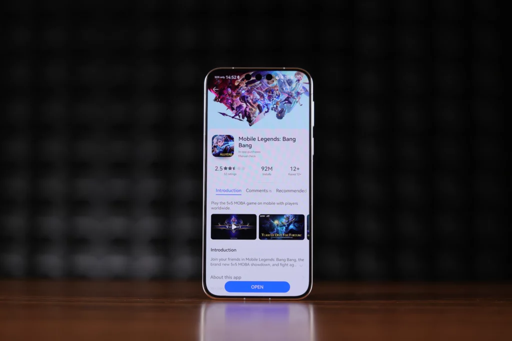 A centered shot of a smartphone standing upright on a wooden table, showing the Mobile Legends: Bang Bang app page inside the Huawei AppGallery. The page shows a 2.5-star rating, 92 million installs, and a blue "OPEN" button at the bottom. The background consists of dark acoustic foam panels.