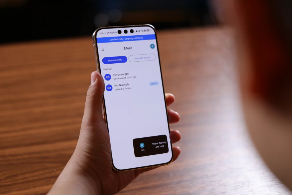 A close-up of a person holding a smartphone displaying the Google Meet app home screen. The interface shows a blue "New meeting" button alongside a history of meeting links, with an active call banner at the very top indicating an ongoing call.