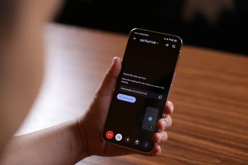 A close-up shot focusing on the back-facing perspective of a person holding a smartphone during an active Google Meet video call. The screen is mostly black, showing meeting details, a "Share invite" link option, and in-call control buttons (mute, video toggle, raise hand) at the bottom.