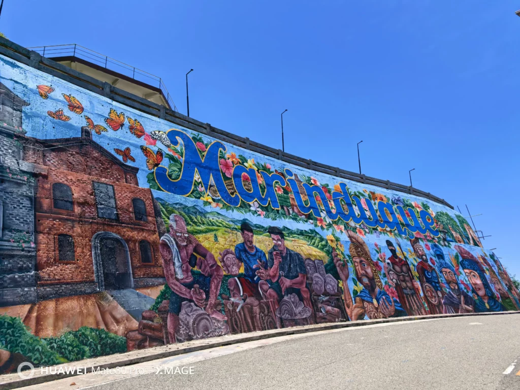 A wide-angle shot of a vibrant, large-scale outdoor mural in Marinduque featuring colorful butterflies, historical buildings, local figures, and traditional masks under a bright blue sky.