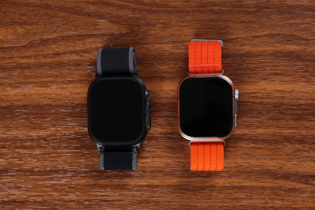 A top-down view of the Apple Watch Ultra 3 (black) and the Huawei Watch Fit 5 Pro (orange) resting side-by-side on a wooden surface with their screens off.