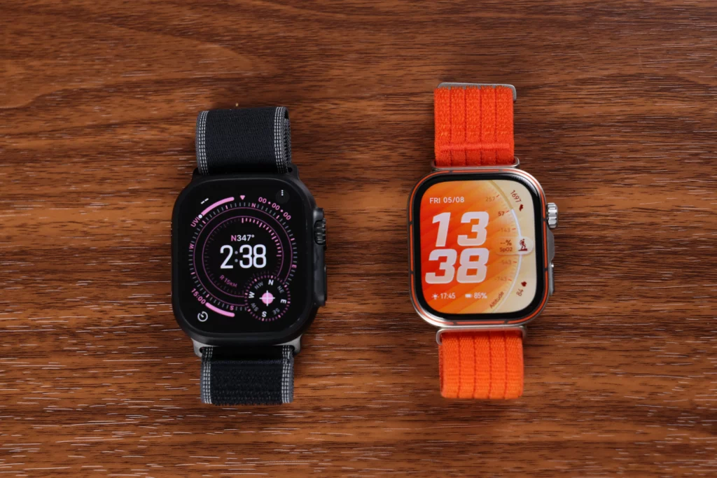 A side-by-side comparison on a wooden table showing the active watch faces of the black Apple Watch Ultra 3 and the orange Huawei Watch Fit 5 Pro.