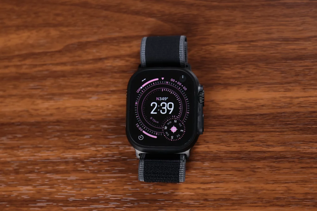 A close-up centered shot of the black Apple Watch Ultra 3 with its compass-style watch face illuminated while resting on a wooden surface.