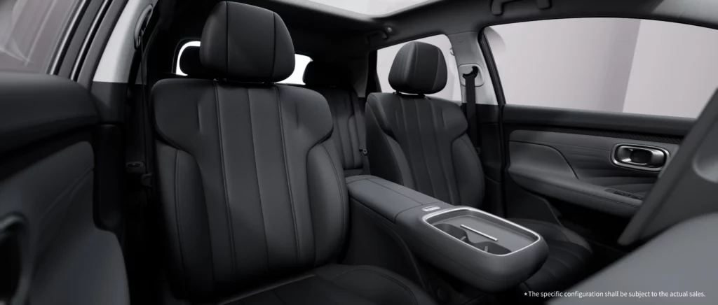 A wide interior view of the GAC AION UT cabin from the front passenger side, highlighting the spacious black leather seats with modern vertical stitching. The center console features a sleek integrated cup holder and armrest, while the panoramic sunroof fills the open, lounge-like cabin with natural light.