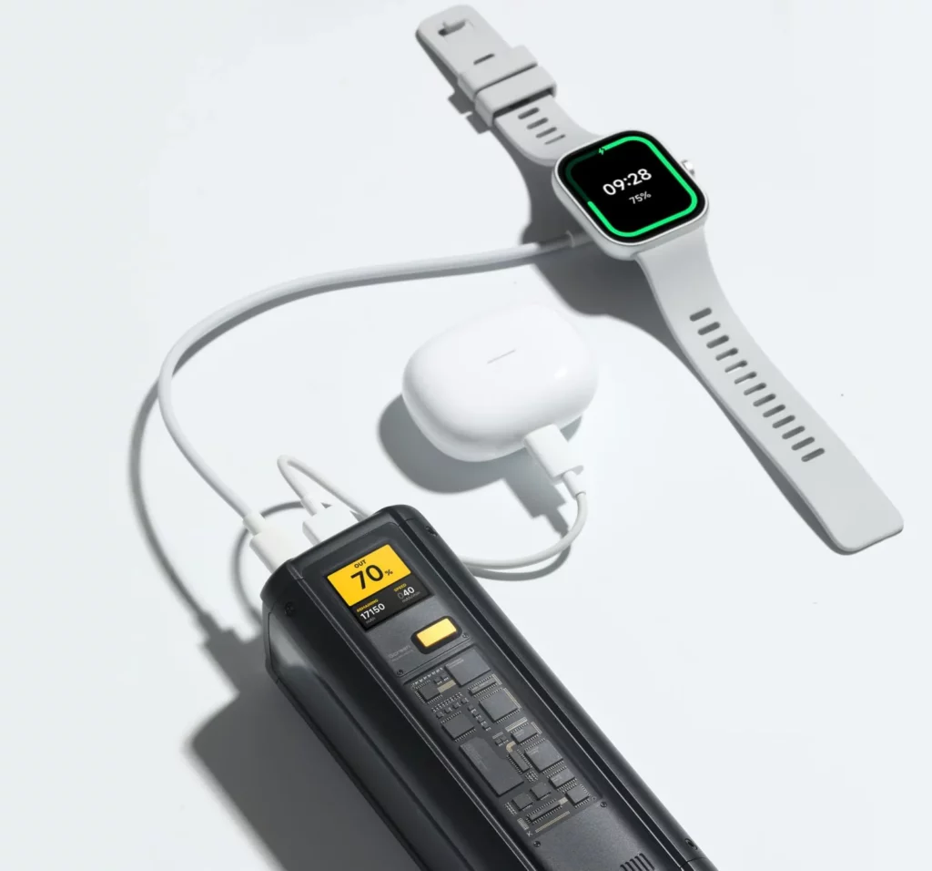 High-angle shot of a black Xiaomi HyperCharge power bank with a transparent casing showing internal circuitry. It is wired to and charging a white smartwatch, showing 75% battery, and a pair of white wireless earbuds on a bright white surface.