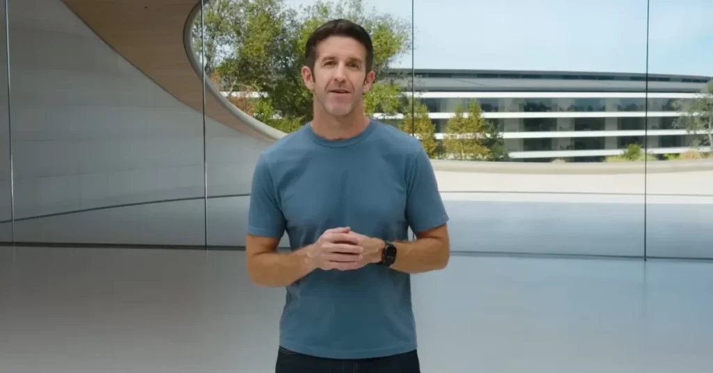 Apple executive John Ternus stands inside a modern, brightly lit glass structure at Apple Park, speaking to the camera with his hands clasped. He is wearing a simple blue t-shirt and a dark Apple Watch, with the curved architecture of the campus visible through the massive glass windows behind him.