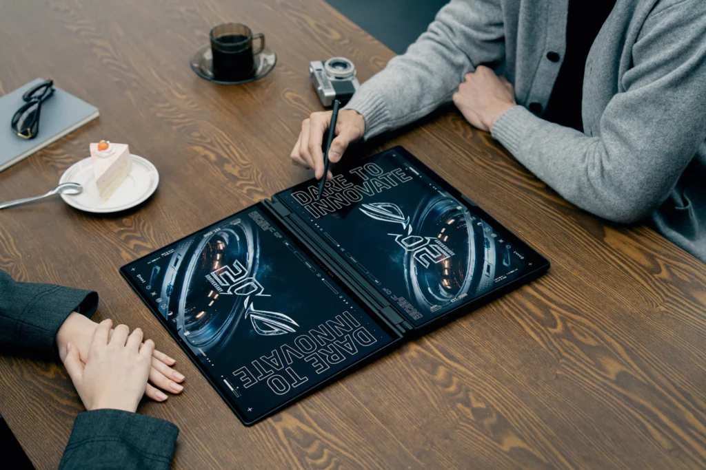 A high-angle shot looking down at a wooden table where two people are collaborating. A dual-screen ASUS ROG Zephyrus laptop is laid flat on the table, showing two large screens displaying matching blue graphic designs with the text "DARE TO INNOVATE," while one person holds a stylus over the screen.