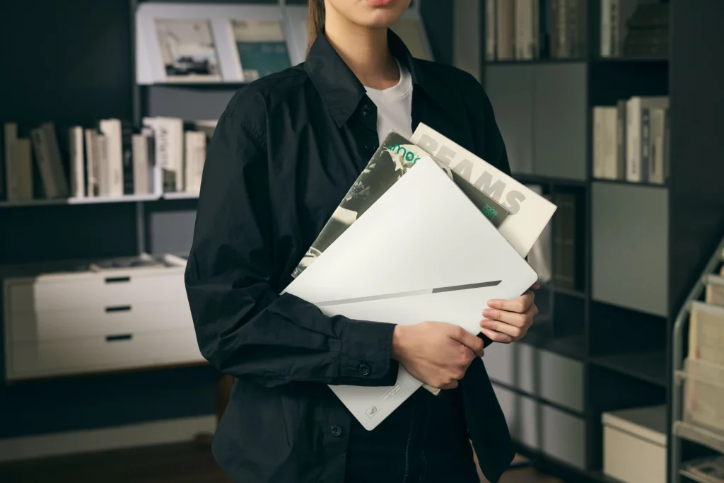 A person in a black button-down shirt holding a sleek, white ASUS ROG Zephyrus G14 laptop under their arm alongside several magazines, with a modern, dark bookshelf filled with books in the background.
