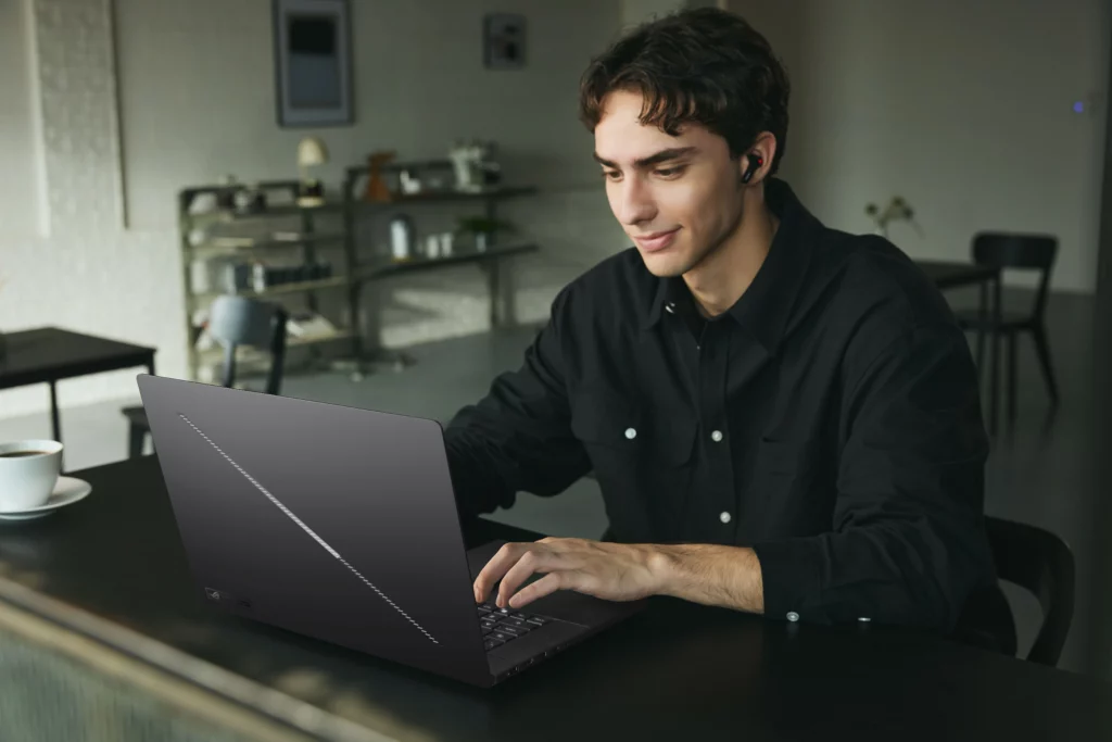 A young man wearing a black shirt and wireless earbuds sitting at a dark table in a cafe, smiling while typing on an ASUS ROG Zephyrus G16 laptop. The laptop lid features a diagonal Slash Lighting strip.