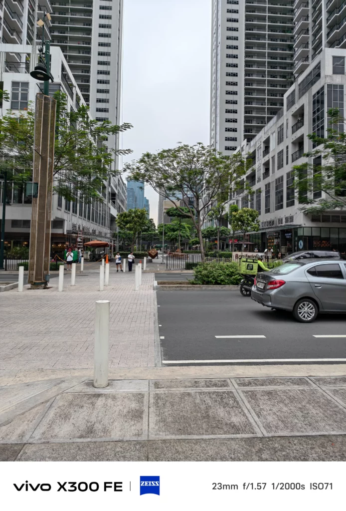 A wide-angle street-level photograph taken in an urban area with tall modern residential buildings lining both sides. The scene features a paved walkway on the left and an asphalt road on the right with a gray car driving by. In the distant center of the street, small figures of pedestrians and distant outdoor decor are barely visible. A white banner at the bottom displays camera specifications: 23mm, f/1.57, 1/2000s, ISO71.