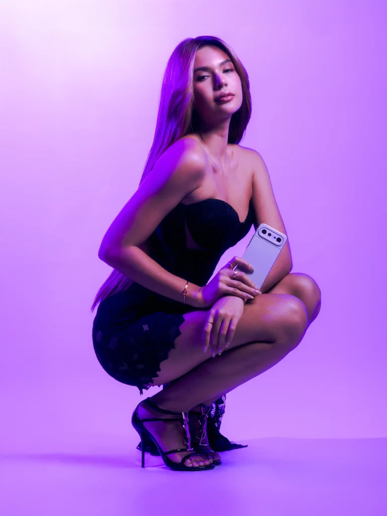 A studio portrait of a woman with long straight hair crouching in a dark black strapless dress against a solid purple background. She holds a light blue vivo X300 FE smartphone across her knee, looking elegantly off-camera under cool purple studio lighting.