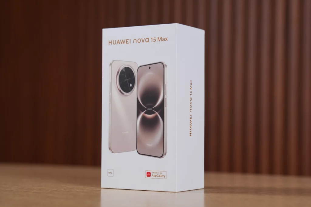 Three-quarter perspective shot of the white retail box for the HUAWEI nova 15 Max smartphone sitting on a wooden table. The front of the box features the product name in gold text and high-quality images of the phone's front and back design. AppGallery and NFC logos are visible near the bottom corner.