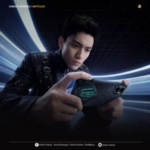 A young man with dark hair wearing a black leather jacket focuses intently while holding a black Infinix gaming smartphone horizontally. The back of the phone features a textured design with glowing green accent lines. The background shows a futuristic, curved tunnel with streaks of blue and white light. The top left corner features the text "UNBOX DIARIES / ARTICLES," and social media handles are listed along the bottom border.