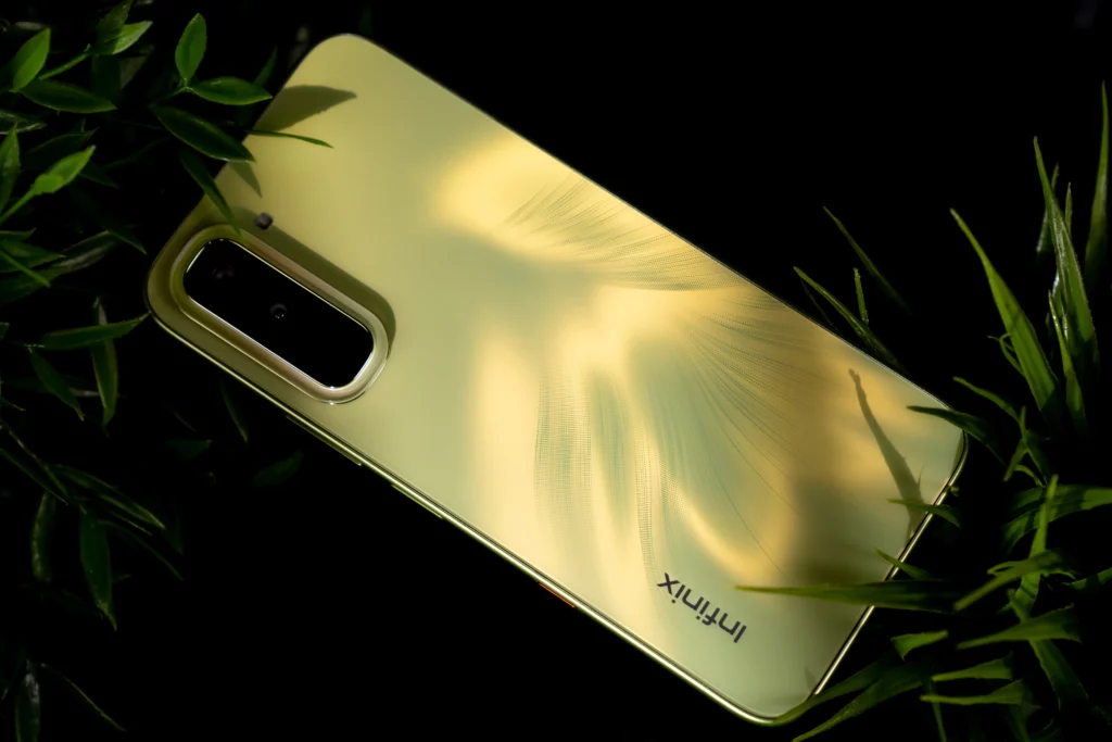 The textured, lime-green back panel of the Infinix HOT 70 smartphone lying on a dark surface, partially framed by green foliage. The dual-camera module is visible at the top left, and light creates a feathered pattern across the glossy finish.