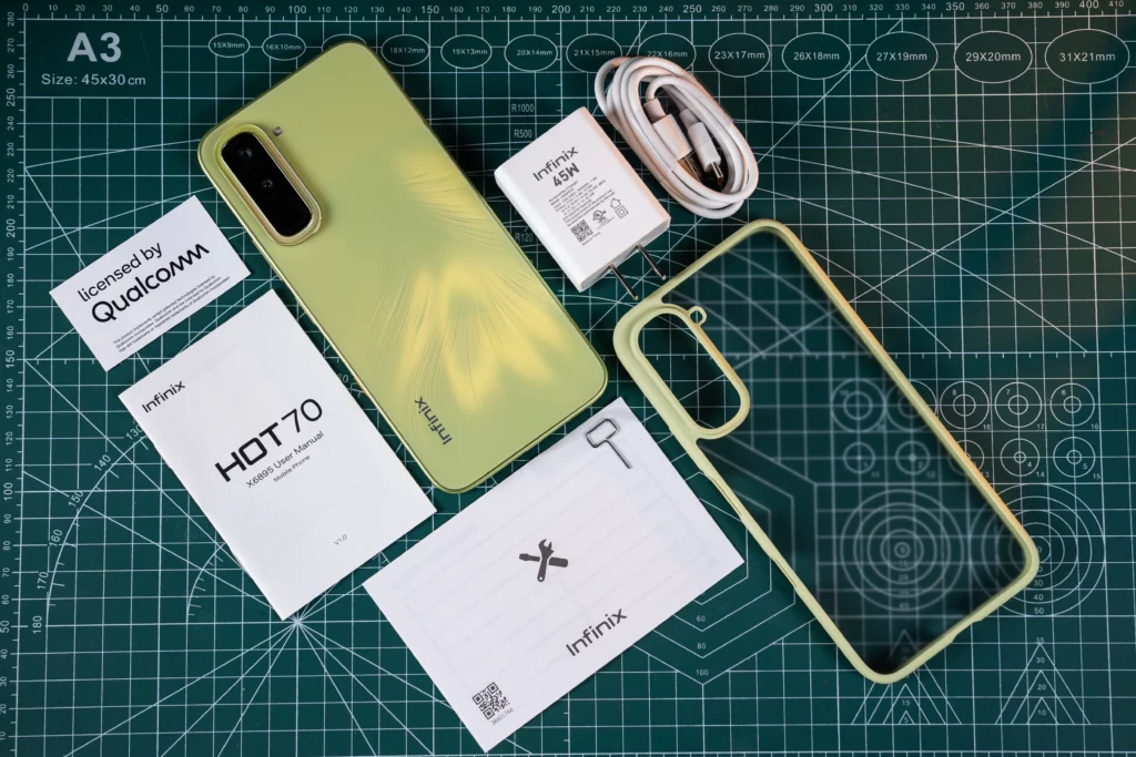 An overhead unboxing shot of the Infinix HOT 70 smartphone and its included accessories arranged on a green grid cutting mat. Items include a 45W charging brick, a white USB cable, a clear phone case with a lime-green border, a user manual, and a SIM ejector tool.