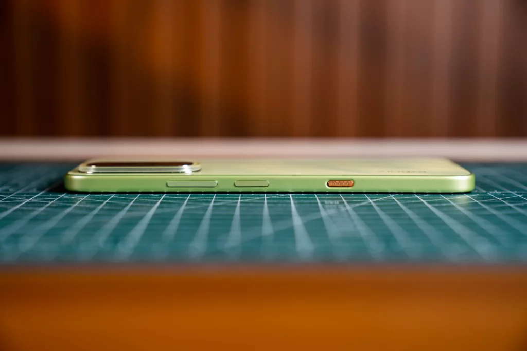 A close-up profile view of the right side of the lime-green Infinix HOT 70 smartphone resting horizontally on a green grid mat. The volume buttons and a textured, copper-colored power button are visible along the slim edge.