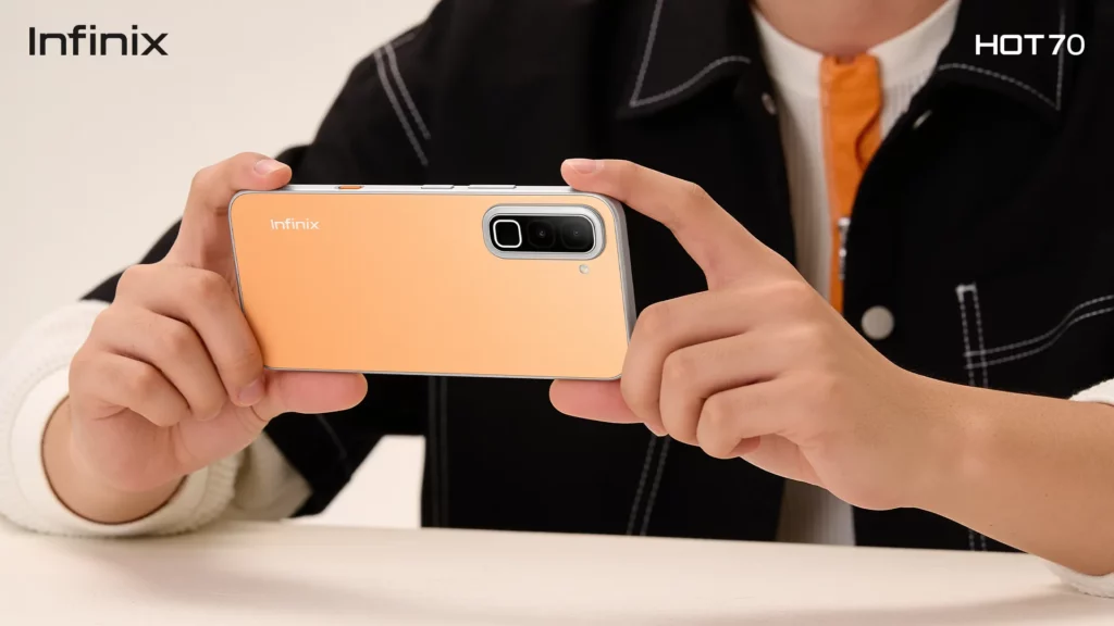 Close-up shot of a person’s hands holding an orange or peach-colored smartphone horizontally as if taking a photo or watching a video. The rear panel of the phone features a clean, minimalist design with the "Infinix" logo on the left and a silver, pill-shaped camera module housing multiple lenses on the right. The person is wearing a dark jacket over an orange and white top. The top corners feature the "Infinix" logo on the left and "HOT 70" on the right.