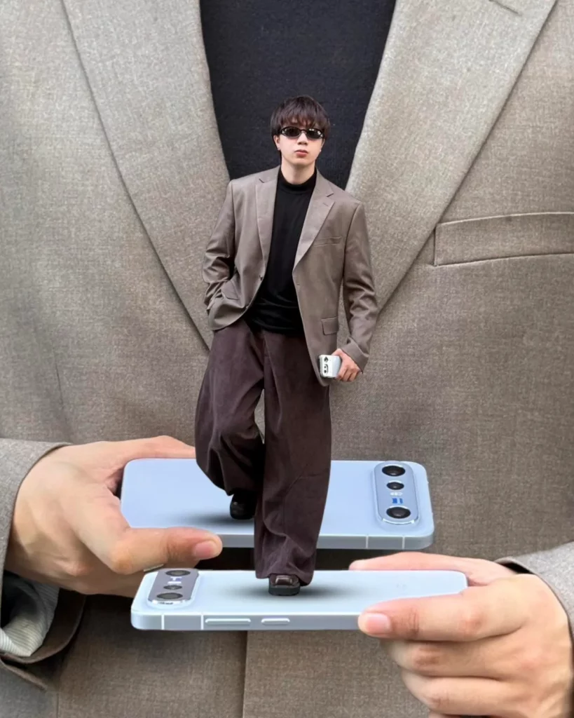 A creative, multi-layered forced perspective photograph featuring a main background of a person wearing a textured brown blazer. In the foreground, two hands hold up two identical light blue vivo X300 FE smartphones horizontally. A miniature-scale full-body image of a stylish young man in sunglasses, a brown blazer, and wide-leg trousers stands directly on top of the lower phone as if it were a platform.