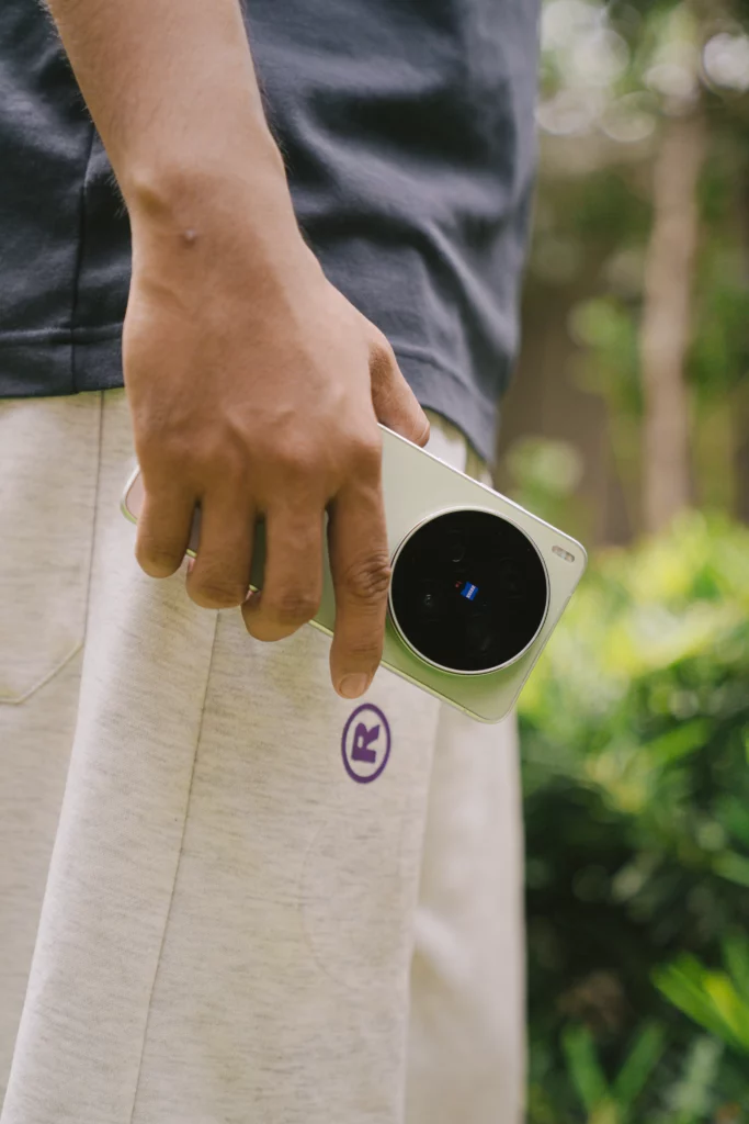 A mid-section shot of a person casually holding a light green vivo X300 Ultra smartphone in one hand by their side. The prominent, circular black rear camera module featuring the blue ZEISS logo is clearly visible against light-colored shorts.