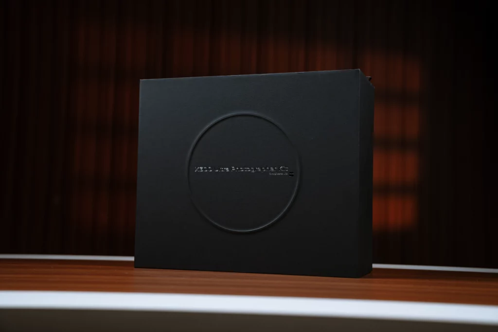 A large, textured matte black presentation box sitting on a wooden surface against a dark background. A large circle is embossed in the center with the silver text "X300 Ultra Photographer Kit Co-engineered with ZEISS" representing an accessory kit for the vivo X300 Ultra.