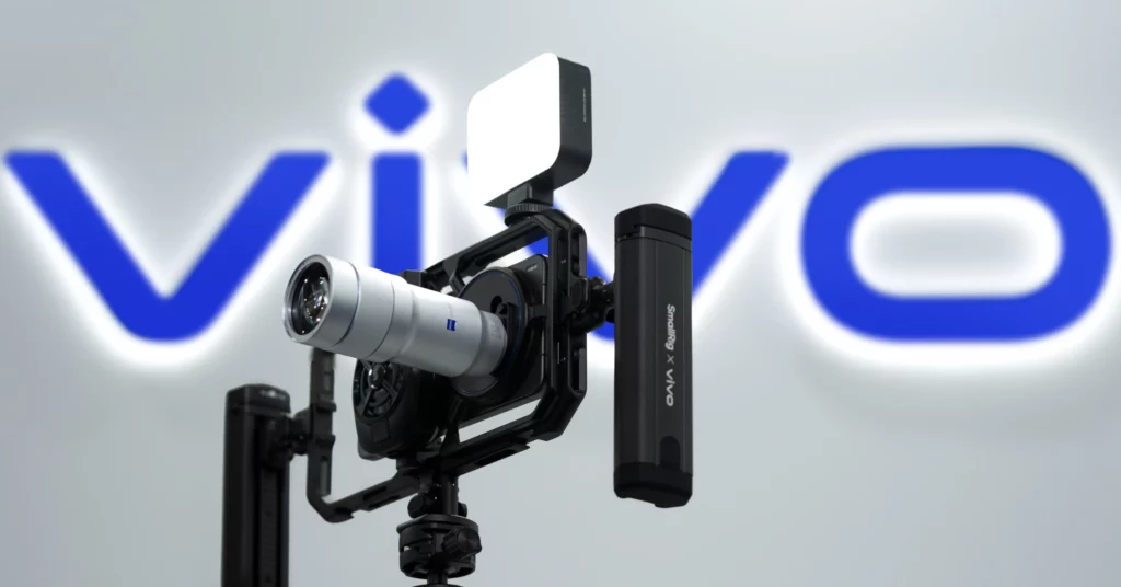 Close-up of a professional mobile filmmaking setup featuring a smartphone secured in a black metal cage rig on a tripod. Attached to the setup are a prominent silver ZEISS lens attachment, an active cooling fan unit behind the phone, a square white LED panel on top, and a side grip handle labeled "SmallRig x vivo". A glowing blue vivo logo is softly blurred in the white background.
