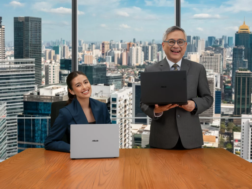 Brand Ambassadors Gabbi Garcia sitting at a wooden conference table with a silver ASUS laptop and Rico Hizon standing happily holding an open black laptop, posing in front of a floor-to-ceiling office window overlooking a cityscape.