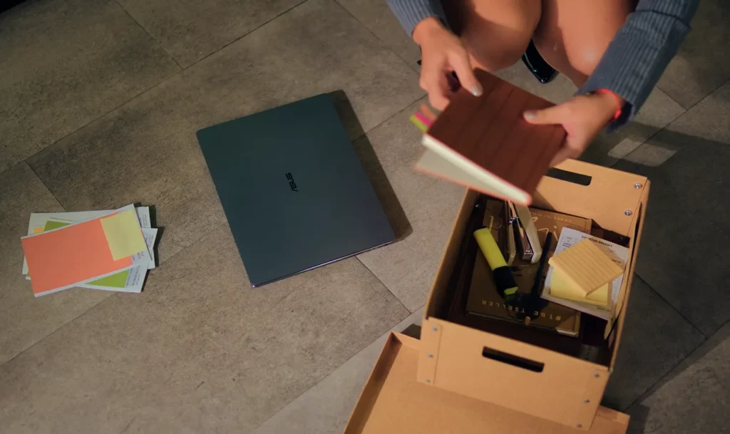 Top-down view of an office floor with a closed gray ASUS ExpertBook Ultra laptop resting next to office supplies and a person packing a cardboard storage box with notebooks.