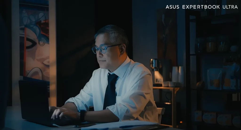 Broadcast journalist Rico Hizon wearing glasses, a shirt, and a tie, typing intently on a dark ASUS ExpertBook Ultra laptop at a desk during late-night hours.