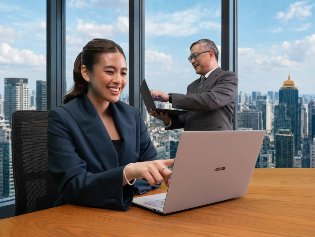 Gabbi Garcia smiling and pointing at her silver ASUS ExpertBook Ultra screen while sitting at a desk, with Rico Hizon working on his laptop in the background against a city skyline view.
