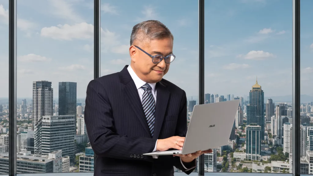 Rico Hizon wearing a navy suit and glasses, smiling as he works on a silver ASUS ExpertBook Ultra while standing in front of a large office window with a panoramic city view.