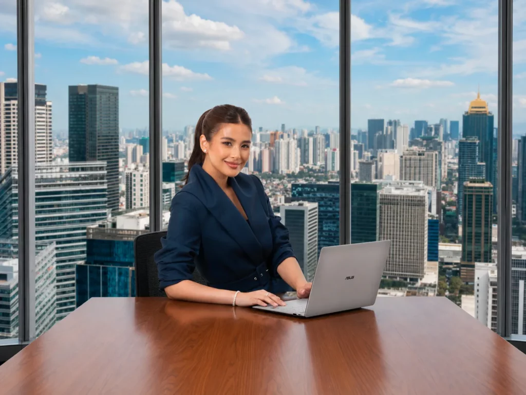Gabbi Garcia dressed in a professional navy blue blazer, smiling at the camera while using a silver ASUS ExpertBook Ultra at a wooden table in an office overlooking high-rise buildings.