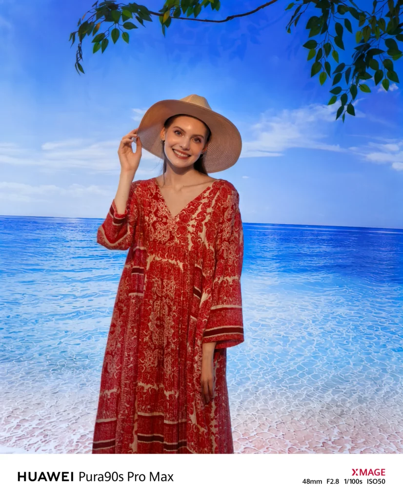 A smiling woman wearing a straw sunhat and a patterned red dress posing against a beach and ocean backdrop.