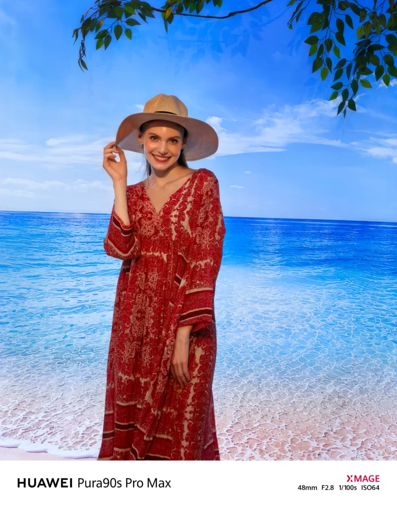 Medium shot of a smiling woman in a sunhat and red patterned dress against a bright blue tropical beach background.