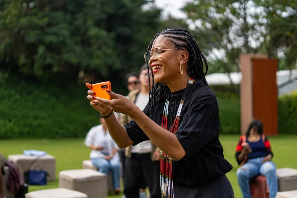 A side-profile medium shot of visual artist Angélica Dass standing outdoors on a green lawn. She is smiling broadly, wearing glasses, a black top, and a vibrant traditional beaded necklace. She is holding up a bright orange TECNO smartphone horizontally with both hands, capturing a photo or video while other event attendees sit blurred in the background.