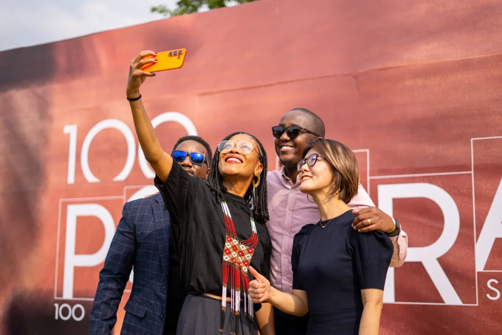 A cheerful group of four diverse people posing closely together for a selfie outdoors. Visual artist Angélica Dass stands in the center holding a bright orange smartphone high in the air to snap the photo. They are smiling warmly, standing in front of a large, rustic-red promotional backdrop featuring the words "100 PORTRAITS".