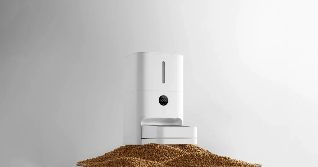 Studio shot of the white Xiaomi Smart Pet Food Feeder 2 with a digital display screen, standing on a large pile of brown kibble against a clean gray background.